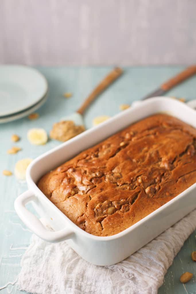Healthy peanut butter banana bread in a loaf pan cooling with bananas in the background.
