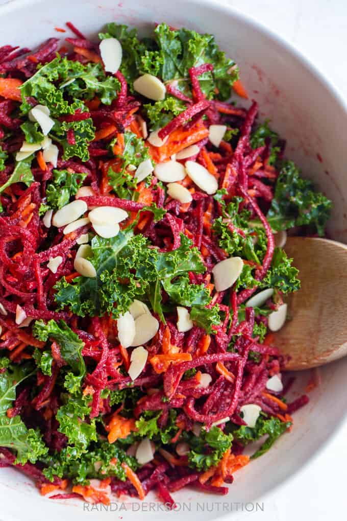 raw beets tossed with shredded carrots and kale with slivered almonds in a white bowl.