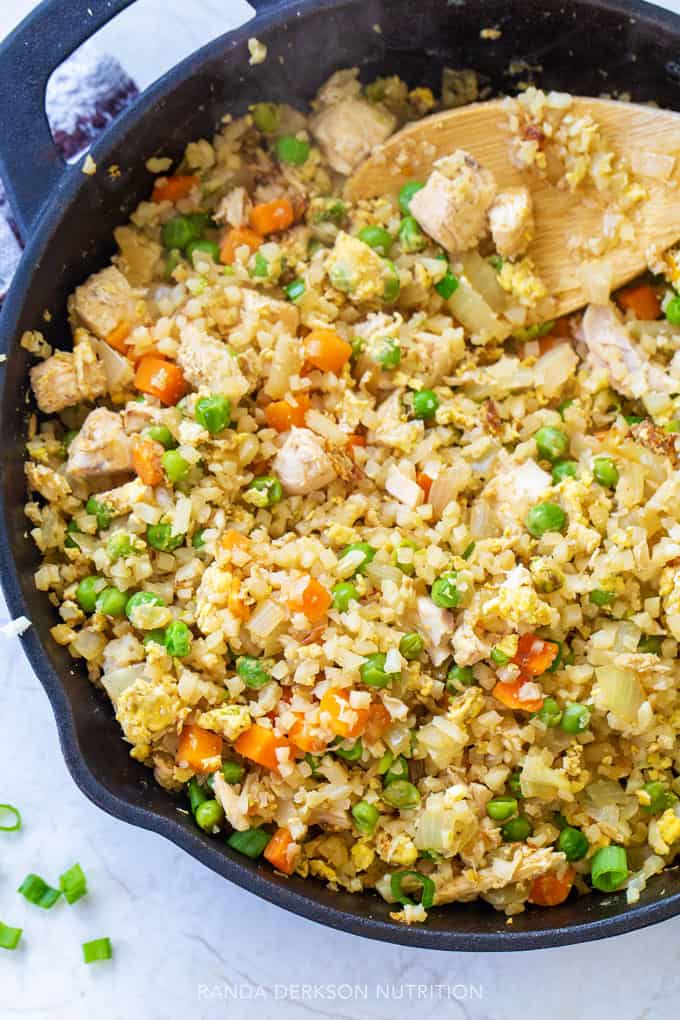 Cauliflower rice, peas, carrots, and chicken being stirred in a cast iron skillet for a healthy dinner.
