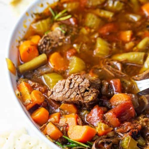 hearty beef and vegetable soup in a white bowl with crackers.