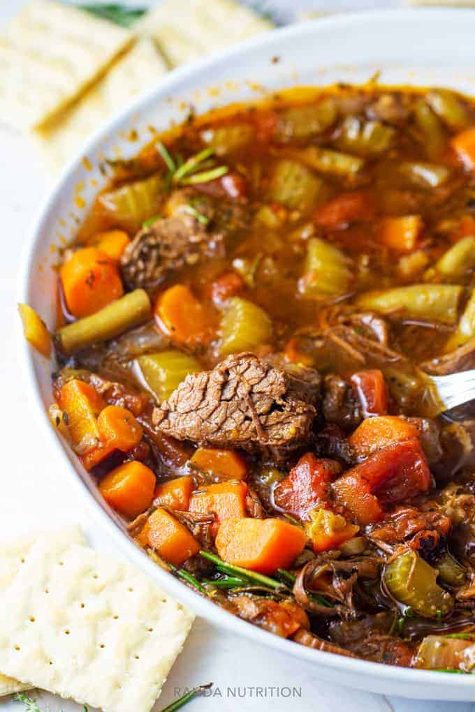 hearty beef and vegetable soup in a white bowl with crackers.