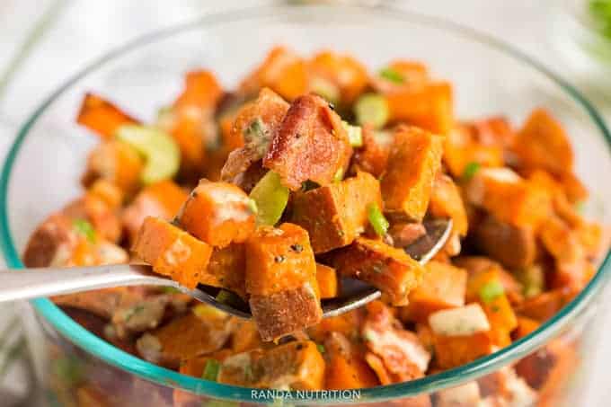 A spoon lifting sweet potato salad with pieces of bacon and a creamy dressing to the camera.