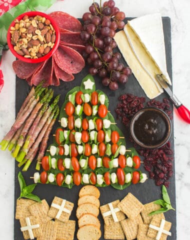 A festive charcuterie board that is simple and easy to make. In shape of a Christmas tree is mozza balls, tomatoes over spinach. Includes a bowl of mixed nuts, grapes, sliced brie cheese, and crackers in the shape of presents.