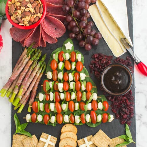 A festive charcuterie board that is simple and easy to make. In shape of a Christmas tree is mozza balls, tomatoes over spinach. Includes a bowl of mixed nuts, grapes, sliced brie cheese, and crackers in the shape of presents.