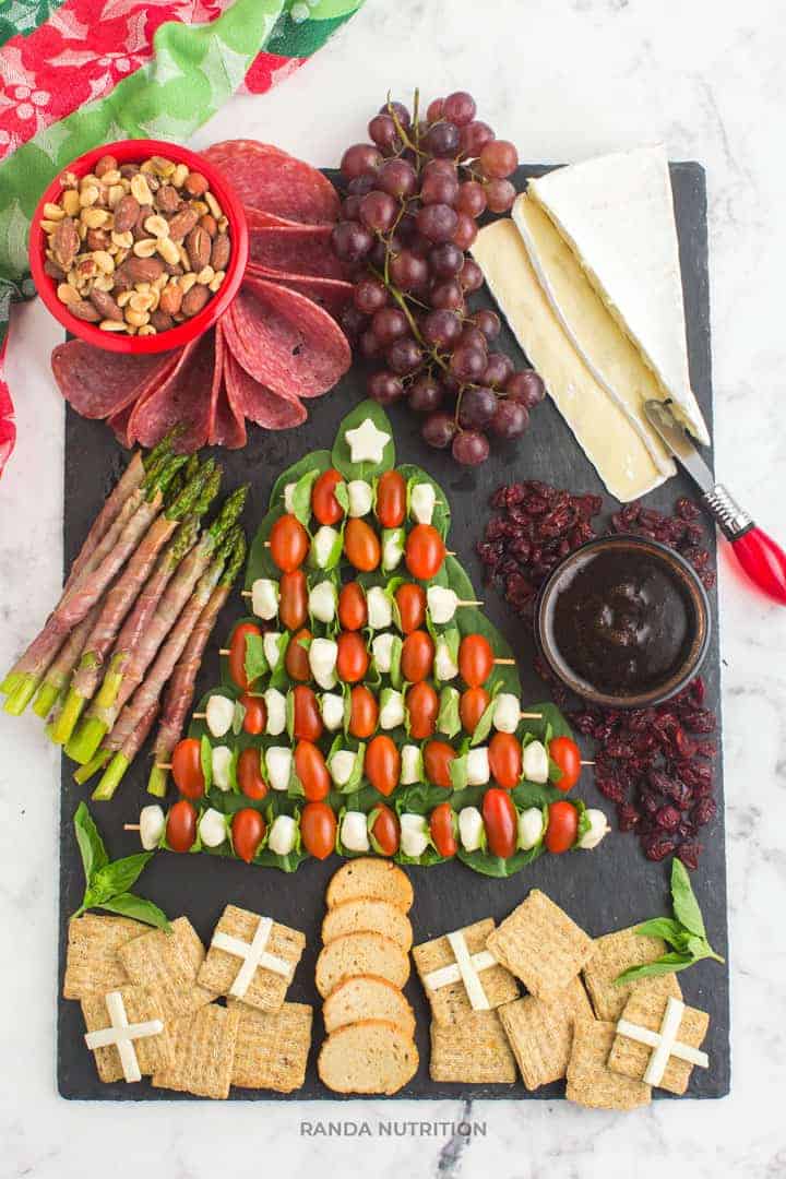 A festive charcuterie board that is simple and easy to make. In shape of a Christmas tree is mozza balls, tomatoes over spinach. Includes a bowl of mixed nuts, grapes, sliced brie cheese, and crackers in the shape of presents.