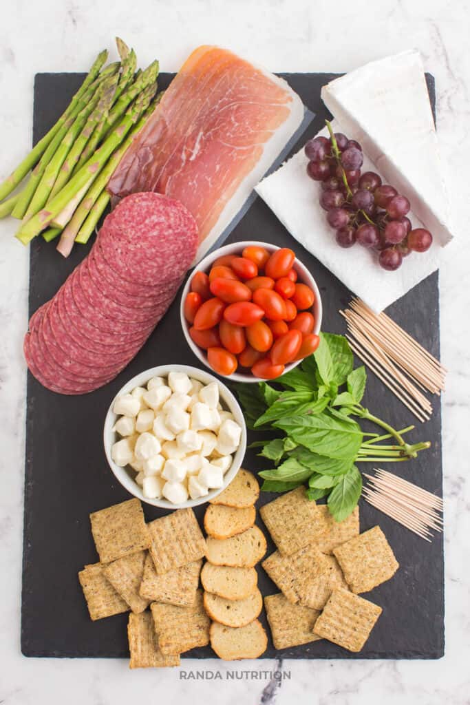 easy charcuterie board ingredients include prosciutto, crackers, balsamic glaze, grapes, herbs, mozza balls, tomatoes, brie, and more