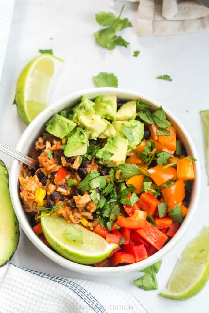 close up overhead view of a white bowl with fresh avocado, orange and red pepper, lime, and cilantro over a rice and chicken bowl