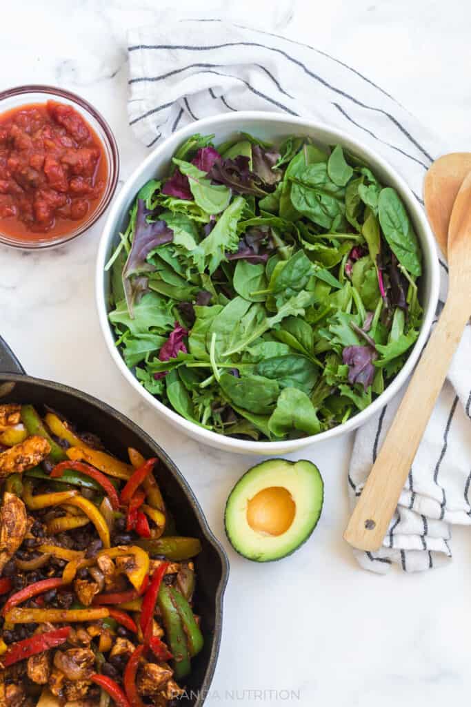 a large bowl of spring mix with a cast iron pan of cooked fajita mix on the side