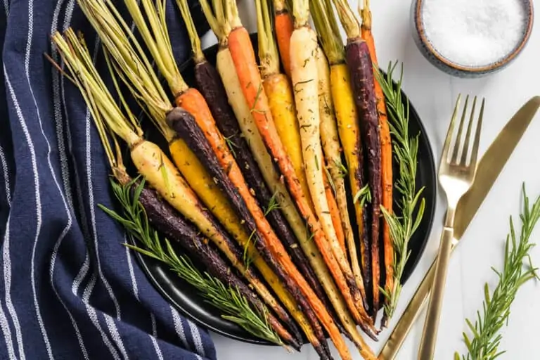 A plate of roasted multicolored carrots garnished with rosemary, placed on a dark plate. Next to the plate are a gold fork and knife, a bowl of salt, and a dark striped cloth.