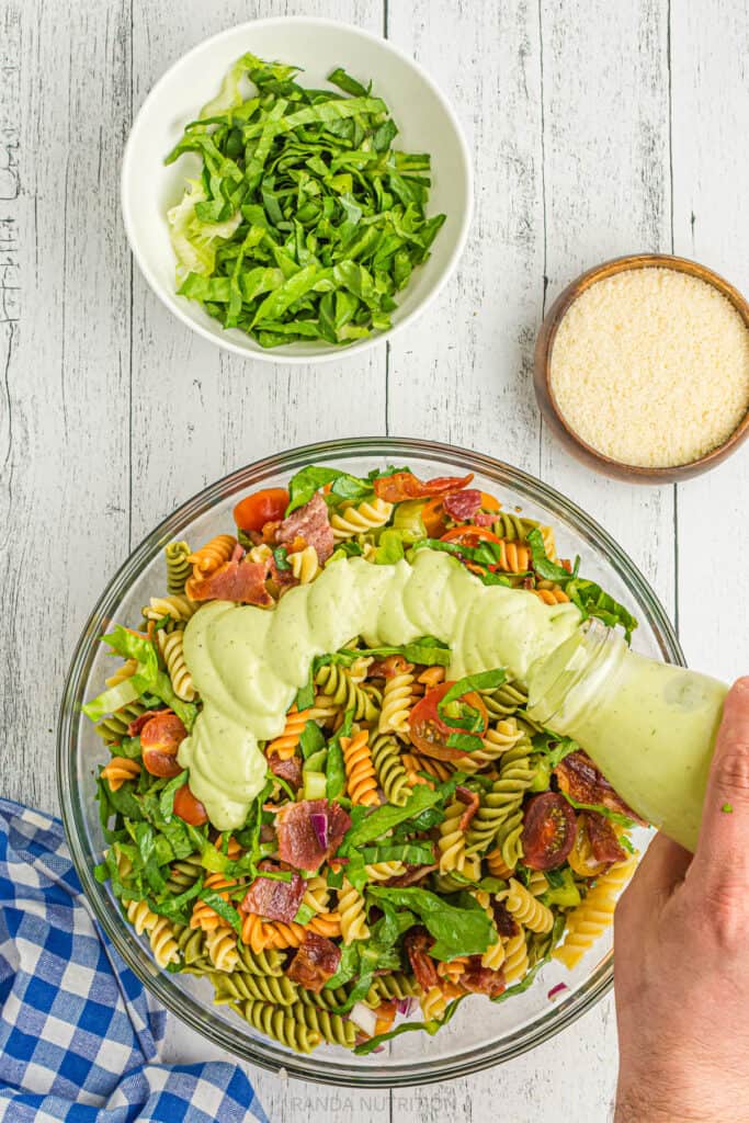 pouring a creamy avocado ranch over pasta salad with bacon lettuce and tomatoes