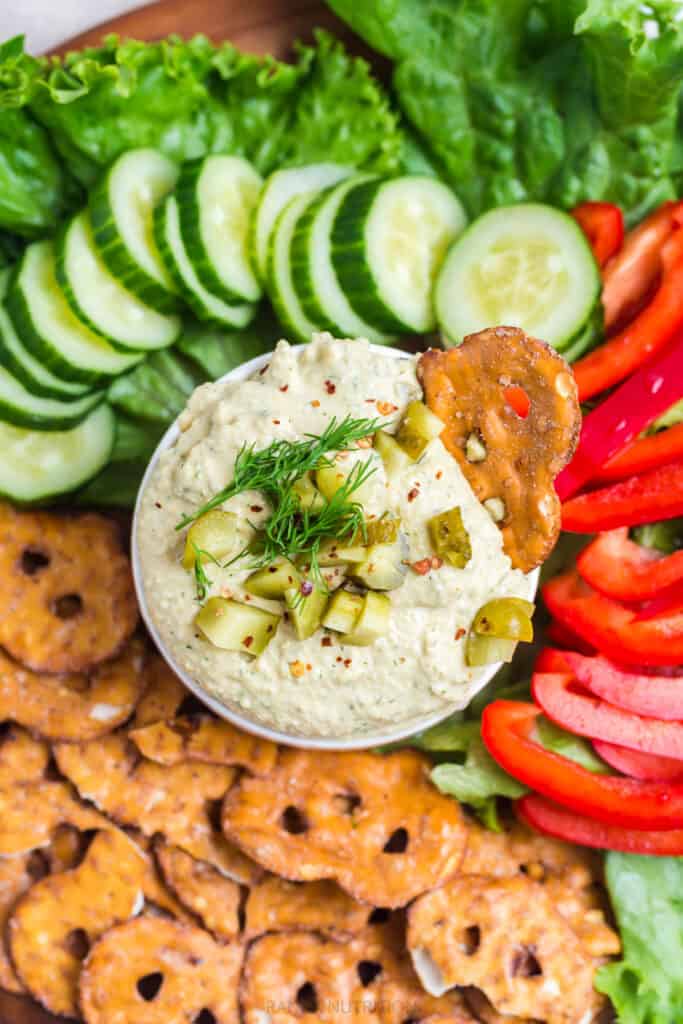 overhead of pickle hummus surrounded by pretzel chips, red peppers, and cucumber slices