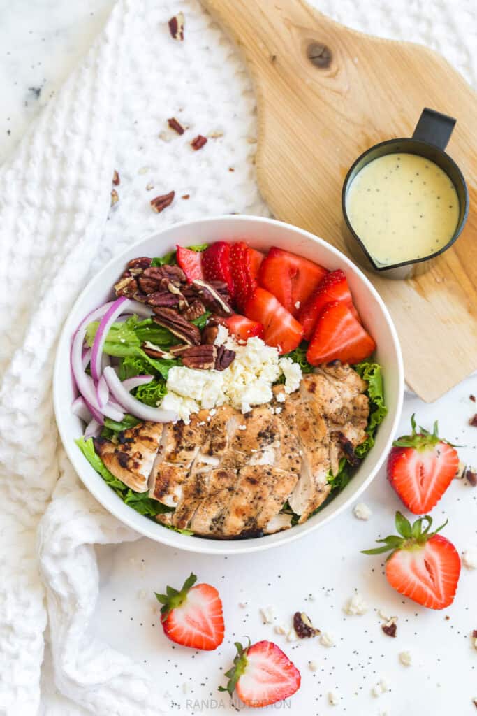 salad bowl with red onion, feta, grilled chicken, pecans, and sliced strawberries with a container of salad dressing on the side